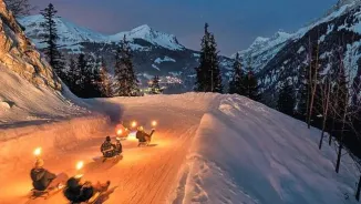 Sledders descending a snowy mountain path at twilight, carrying glowing torches on their sleds.