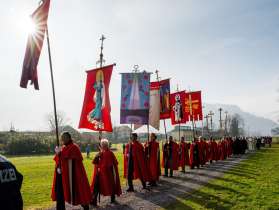 A line of people in red capes carrying ornate religious banners across a field.