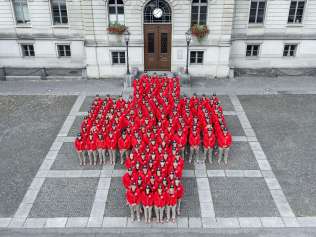 A large group of people wearing red jackets stands arranged in the shape of the Swiss cross in front of a historic building. The formation represents the organizing committee of the ESAF (Swiss Wrestling and Alpine Festival).