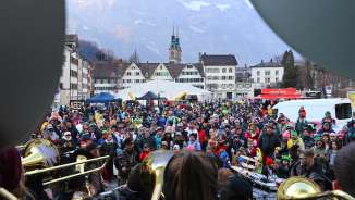 The picture is taken behind/ in the view of a band/ carnival group playing art a carnival in Glarus with people, childen and families watching.
