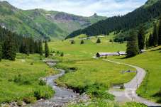 Wide green alpine valley with a stream, hiking paths, scattered farm buildings and forested slopes beneath a blue sky.