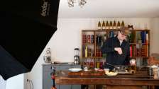 Noah Bachofen standing at a rustic wooden workbench in a kitchen, seasoning a dish in a pan while being lit by professional studio equipment.