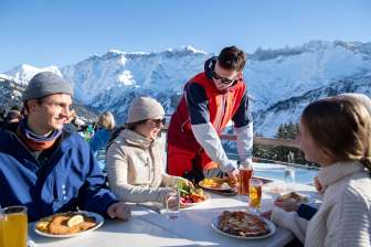 Gäste geniessen Essen auf sonniger Terrasse mit Blick auf verschneite Alpen