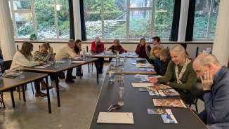 Participants sit at tables during a workshop or presentation at the vernissage of a learning arrangement. Printed materials, photographs, and brochures are spread out as the group discusses and reviews educational content in a bright room.